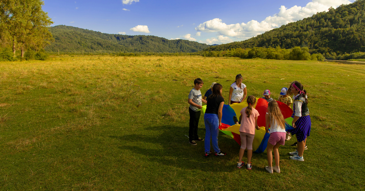 Group of people playing with a colorful parachute in a grassy field with mountains in the background